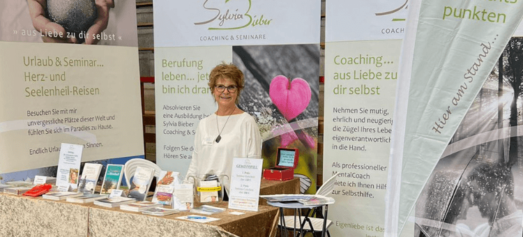 Schirner Tage: Sylvia in ihrem Messestand hinter Büchern und CDs auf dem Tisch und vor den Messewänden