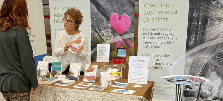 Sylvia im Messestand erklärt einer Besucherin etwas über ihr Buch